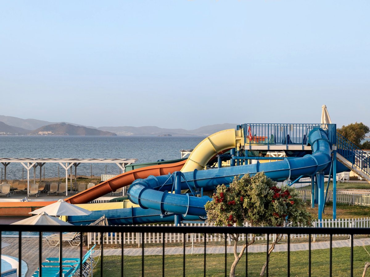 Blue, orange and yellow water slides winding down from a raised platform beside the pool, with the sea and distant hills behind.