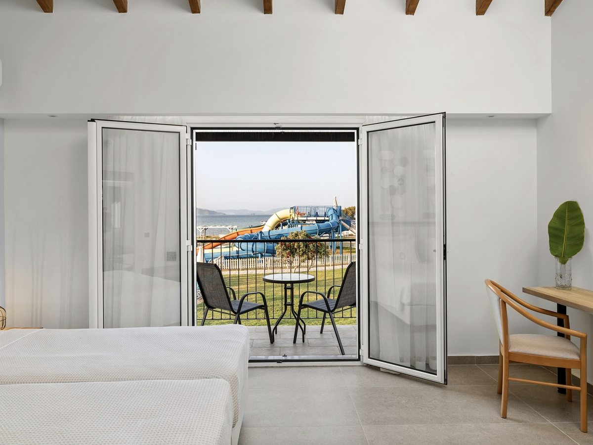 Twin room with open doors onto a balcony with two chairs and a table, looking across a lawn to the blue, orange and yellow water slides and the sea.