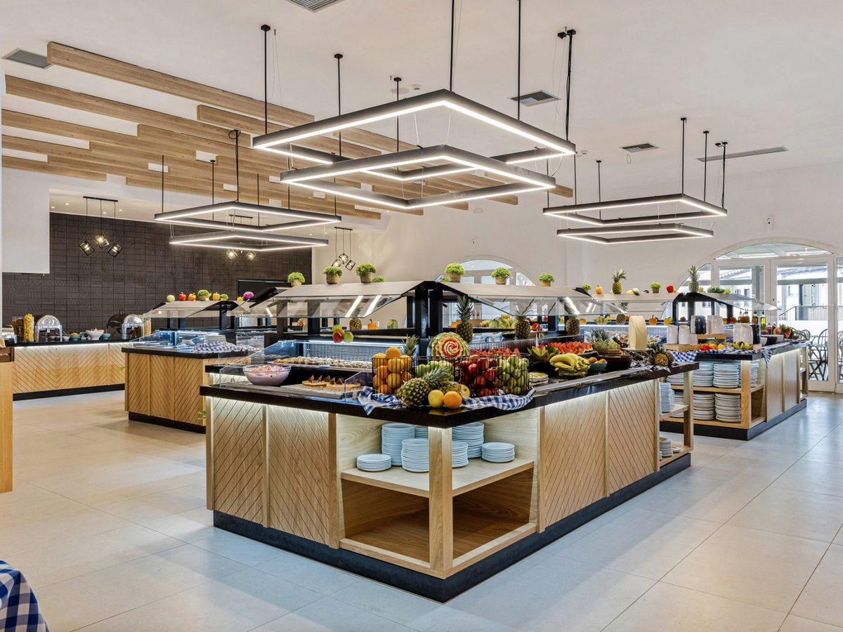 Buffet islands with a fresh fruit display, stacked plates and glass-fronted hot counters under linear pendant lights in a pale, wood-trimmed dining room.
