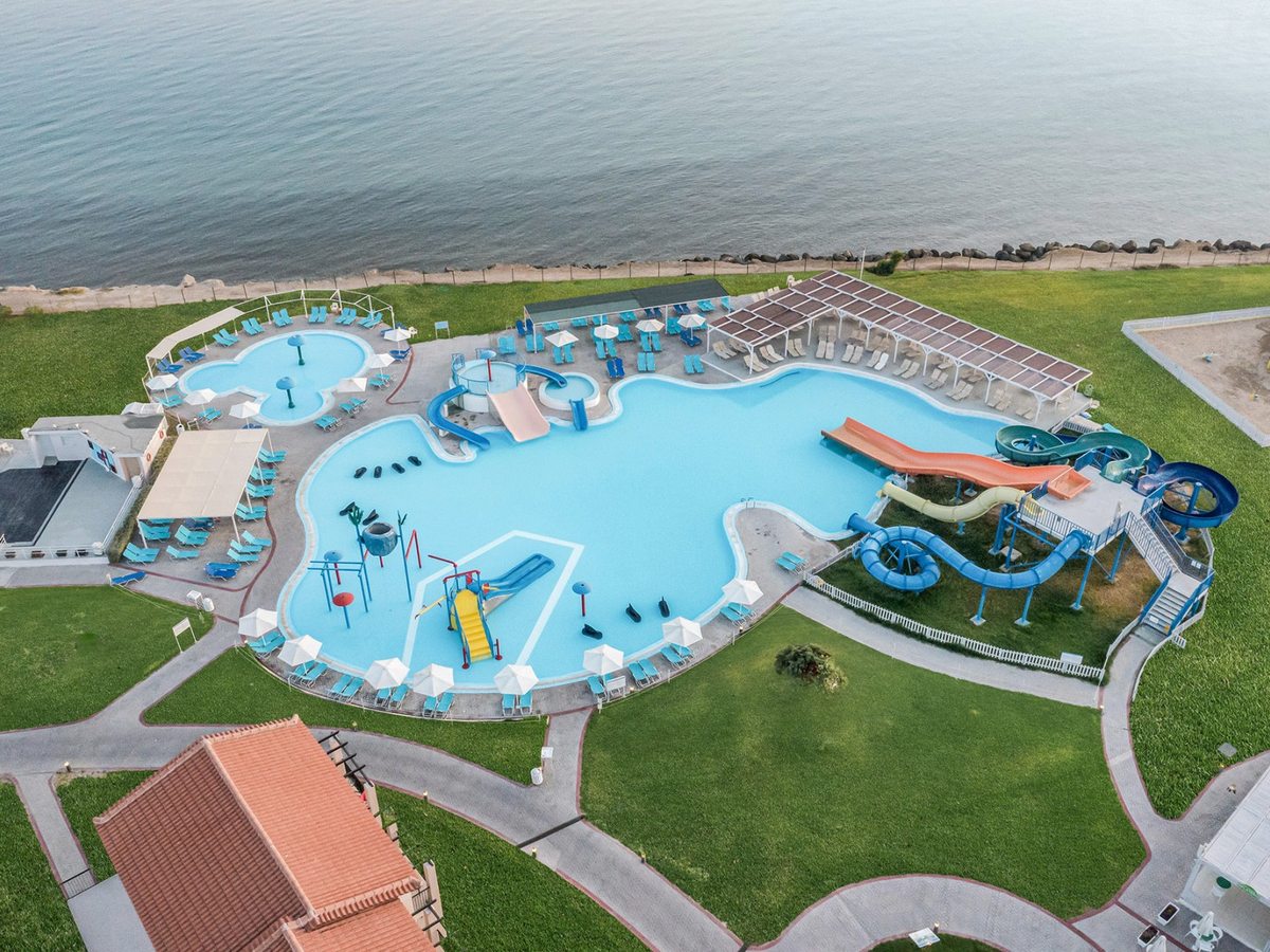Aerial view of the freeform aquapark pool with play structures and a small slide, the cluster of water slides alongside, a separate small round pool, and rows of loungers and parasols by the shore.