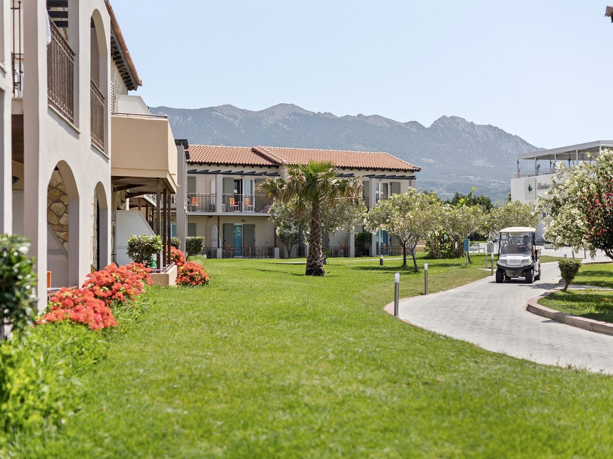 White electric golf buggy on a paved road curving between lawns, flowering shrubs and two-storey room blocks, with mountains behind.