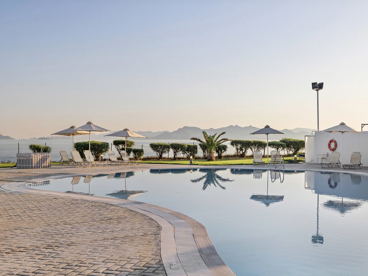Curved pool with a paved surround, sun loungers and parasols, a lifebuoy on a white wall, clipped hedges and the sea and mountains beyond.