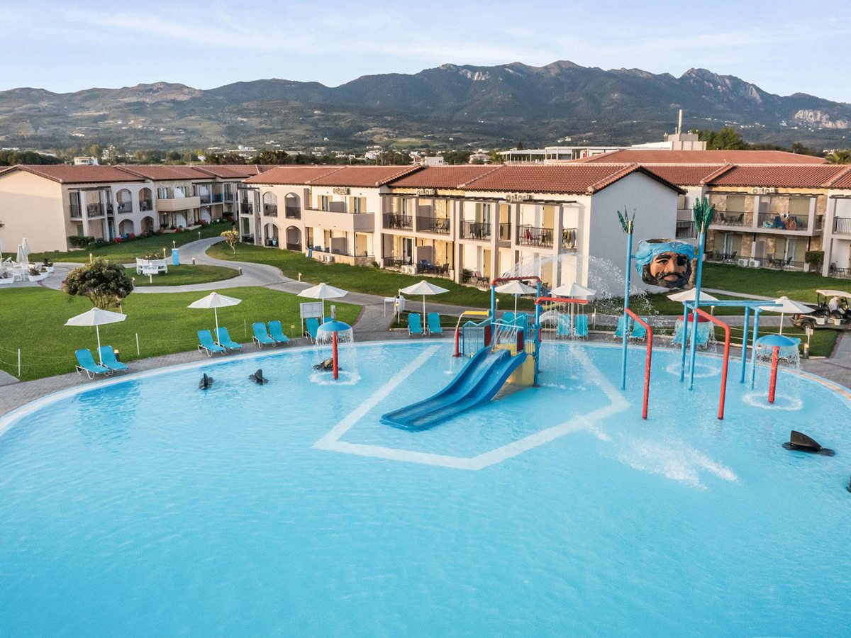 Shallow turquoise pool with a small blue slide, red and blue spray posts, tipping water features and a pirate head sculpture, with room blocks and mountains behind.