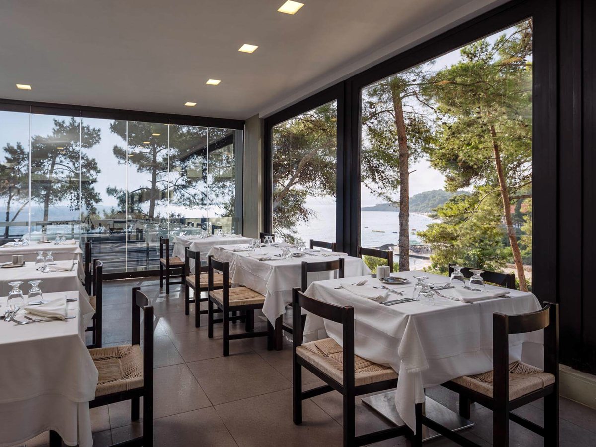Restaurant interior with white-clothed tables and dark wooden chairs, and glass walls onto pine trees and the beach below.