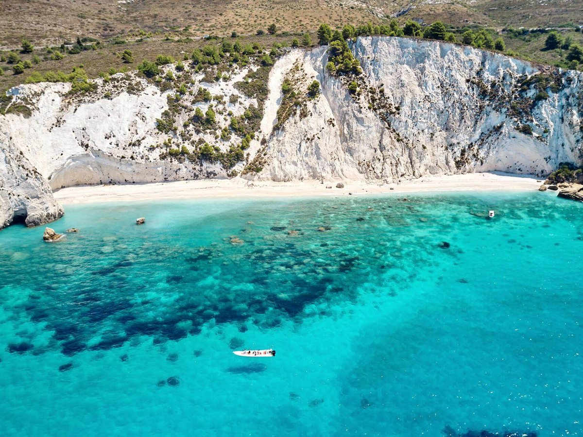 Strip of pale sand beneath tall white cliffs, with clear turquoise water and a small boat anchored offshore.