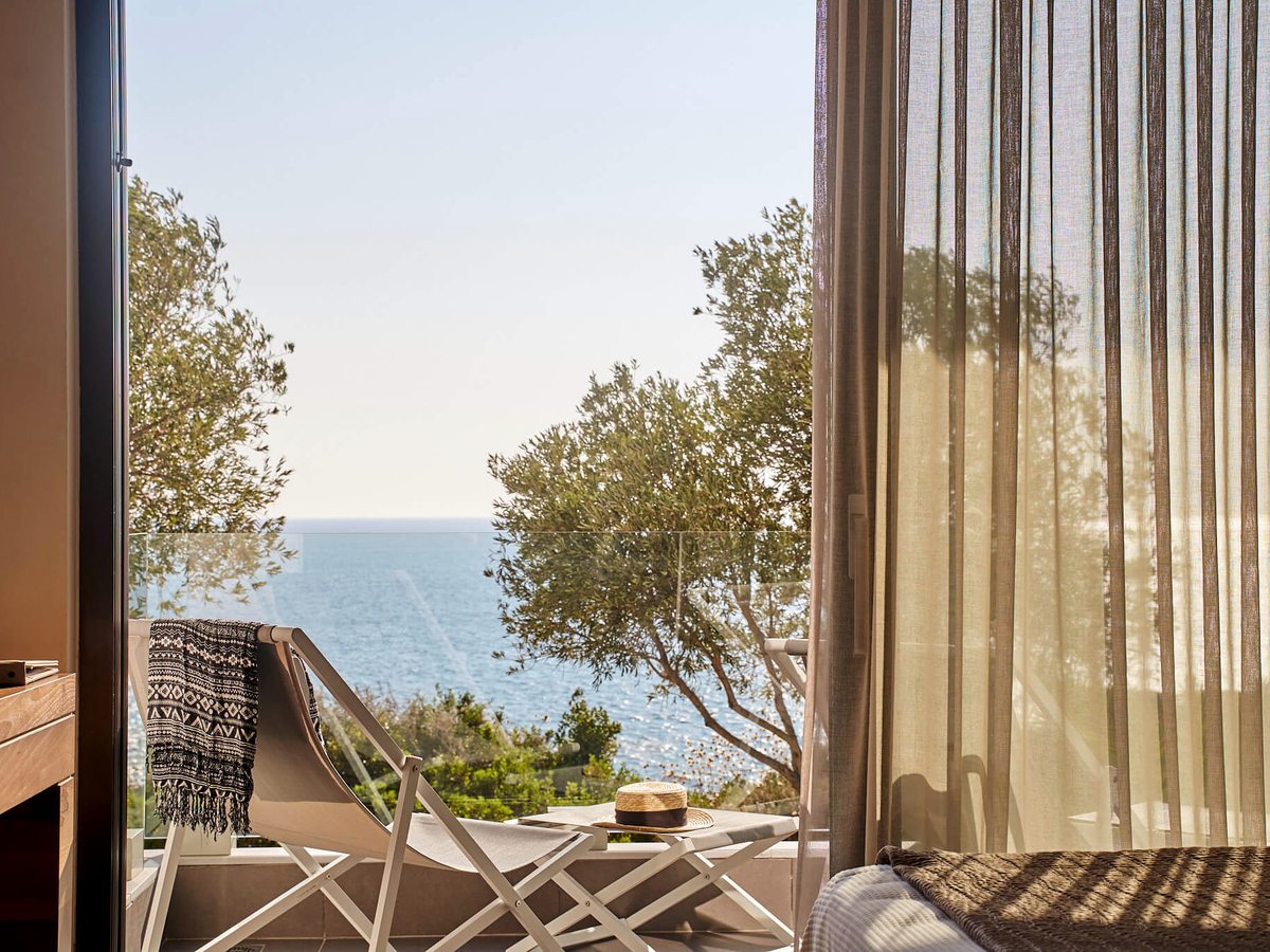 Balcony with two folding chairs and a small table beside an open bedroom door, with olive branches and the sea beyond the glass balustrade.