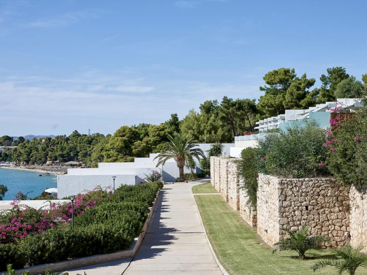 Paved path running between clipped shrubs and a dry-stone wall, with white stepped bungalow terraces on one side and the bay below on the other.