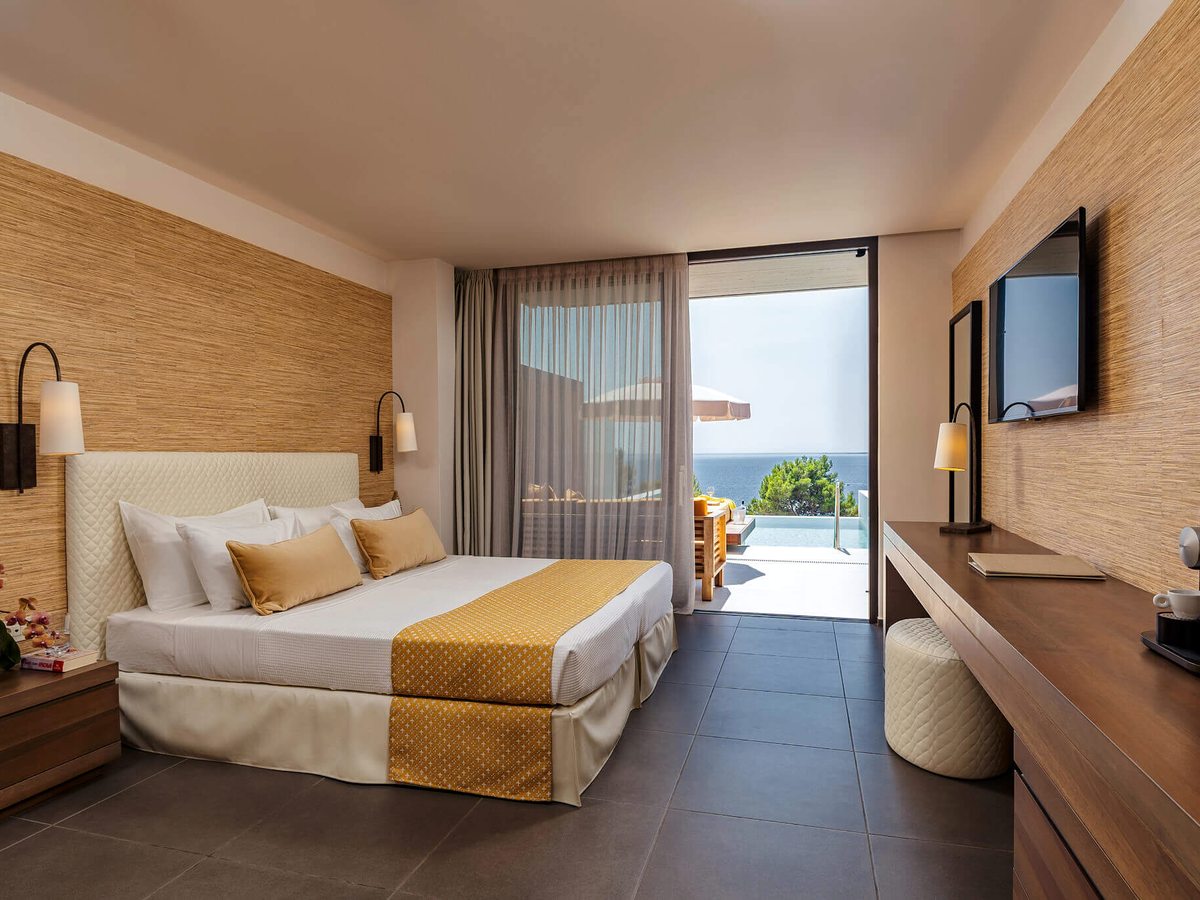 Room with a king bed, woven wall covering and a wooden desk, with sliding doors open onto a terrace with a yellow daybed and a private infinity pool.