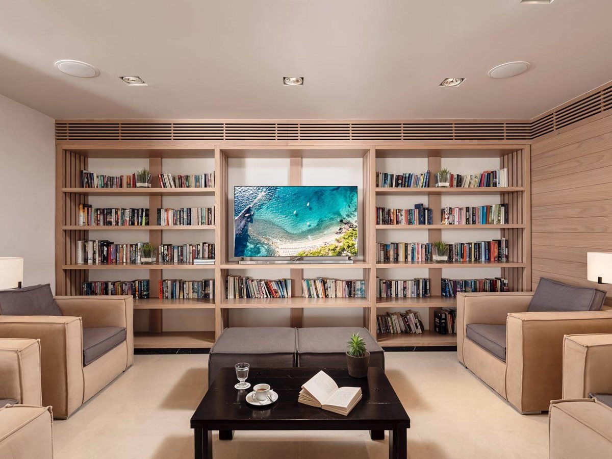 Lounge with pale wooden bookshelves filling one wall around a screen, with beige armchairs and a dark coffee table.