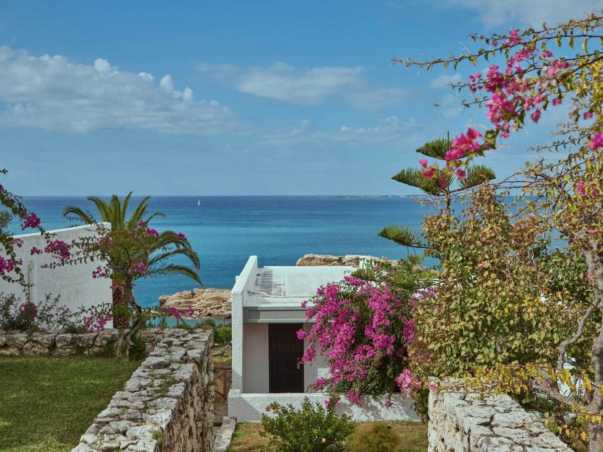 Pink bougainvillea flowering over a small white building, with lawns, dry-stone walls, palms and the blue Ionian Sea beyond.