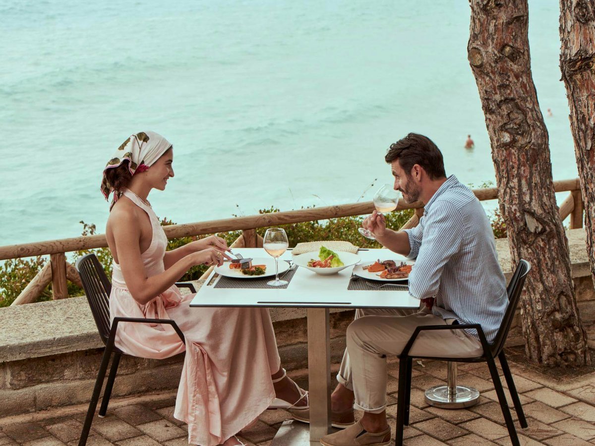 Two people eating lunch at a white table on a paved terrace behind a wooden rail, with the sea just below and pine trunks alongside.