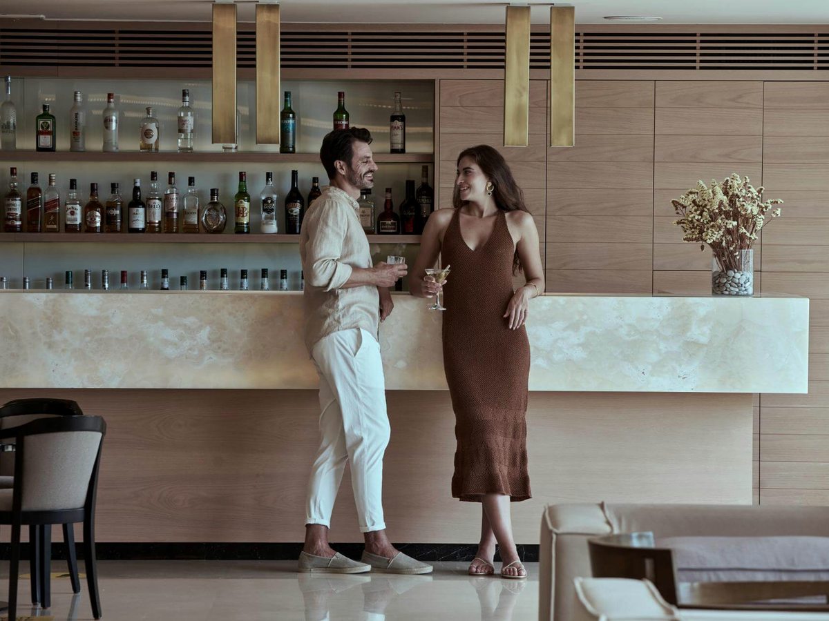Long pale onyx bar counter with brass pendant lights and shelves of spirits behind, and two guests standing with drinks.