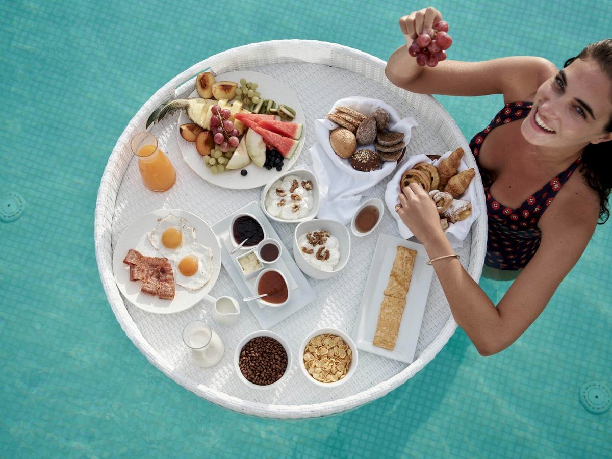 Round white tray floating on turquoise water, loaded with eggs, bacon, pastries, fruit, yoghurt and juice, with a swimmer alongside.