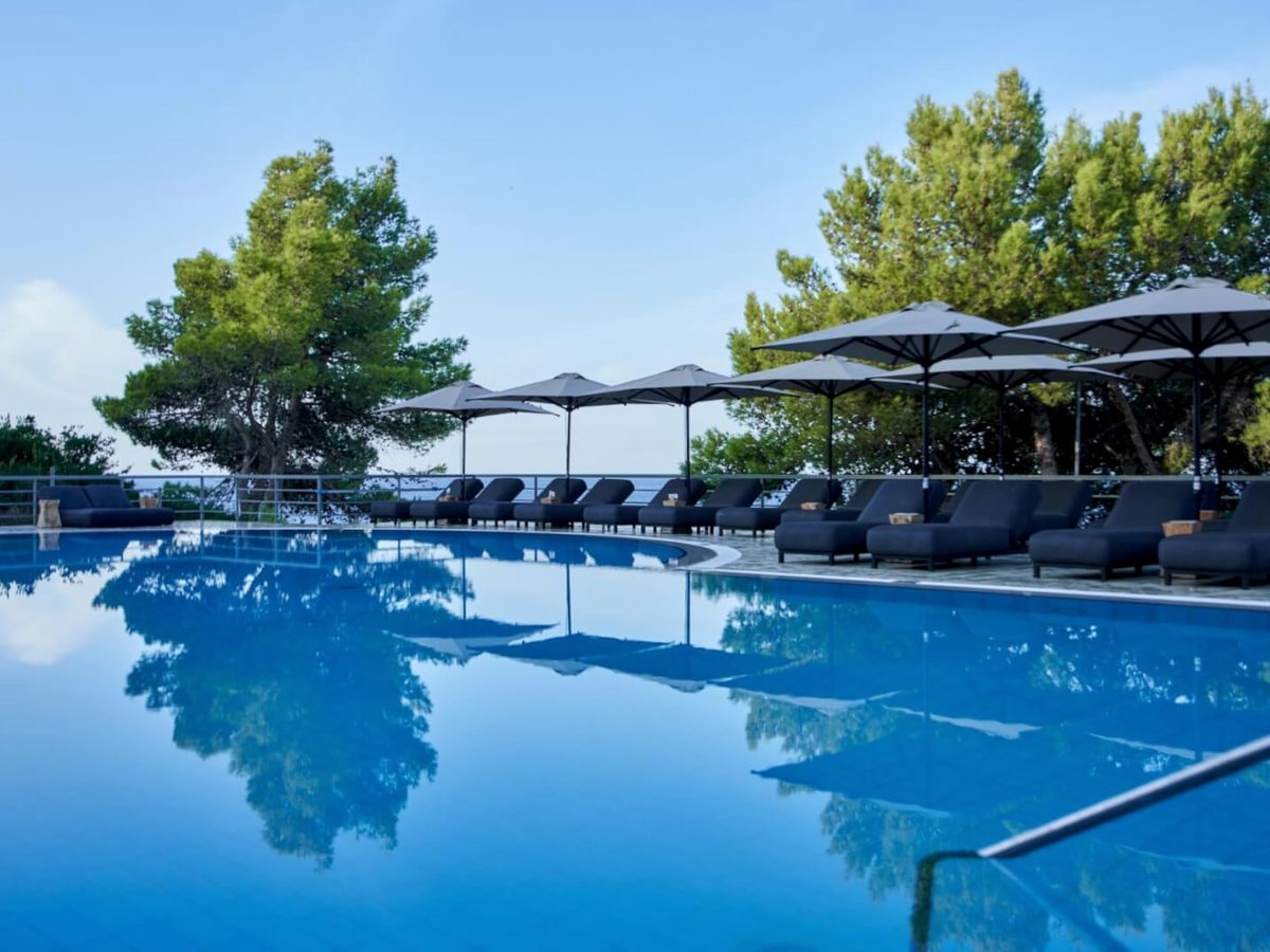 Large pool with a rounded end, dark sun loungers and parasols along one side and pine trees behind, under a clear sky.