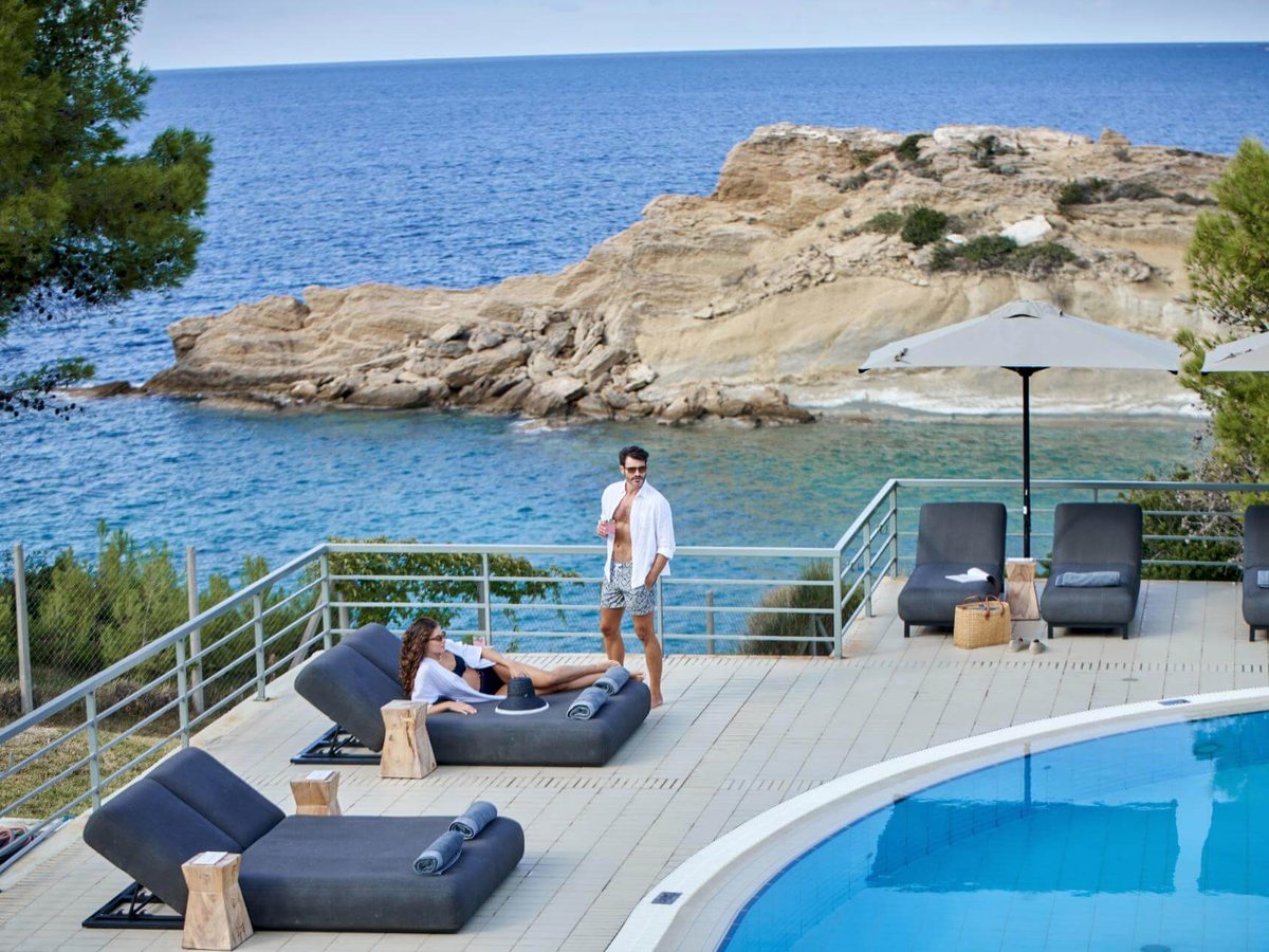 Pool terrace with dark sun loungers and a parasol beside the water, a railing at the cliff edge and a rocky headland in the sea beyond.