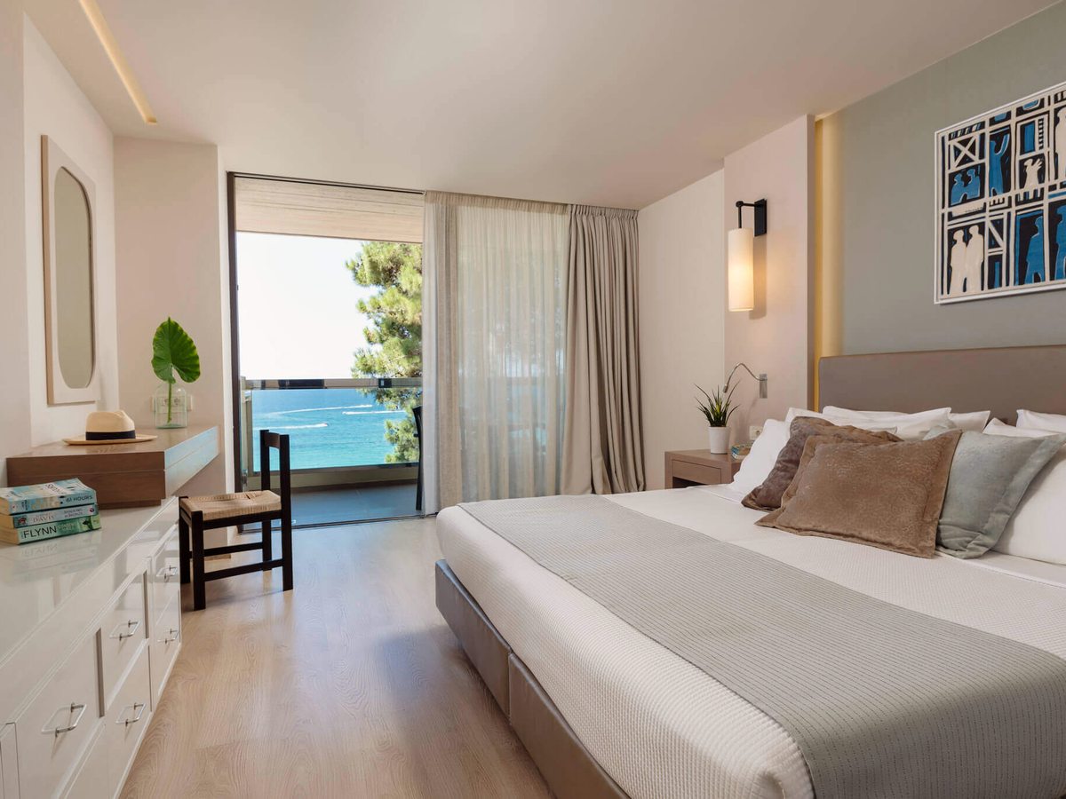 Hotel room with a king bed, a white dresser and a mirror, and sliding doors onto a balcony framed by pine trees above the sea.