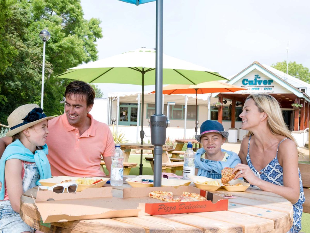 A family of four eating pizza and chips at a round wooden picnic table under a green parasol, with the Culver building behind.