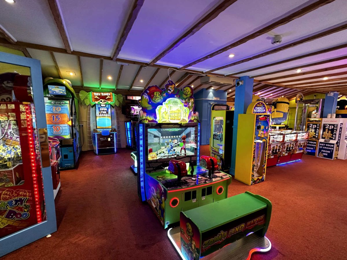Rows of illuminated arcade cabinets, claw machines and coin pushers in a red-carpeted room with a beamed ceiling lit purple.