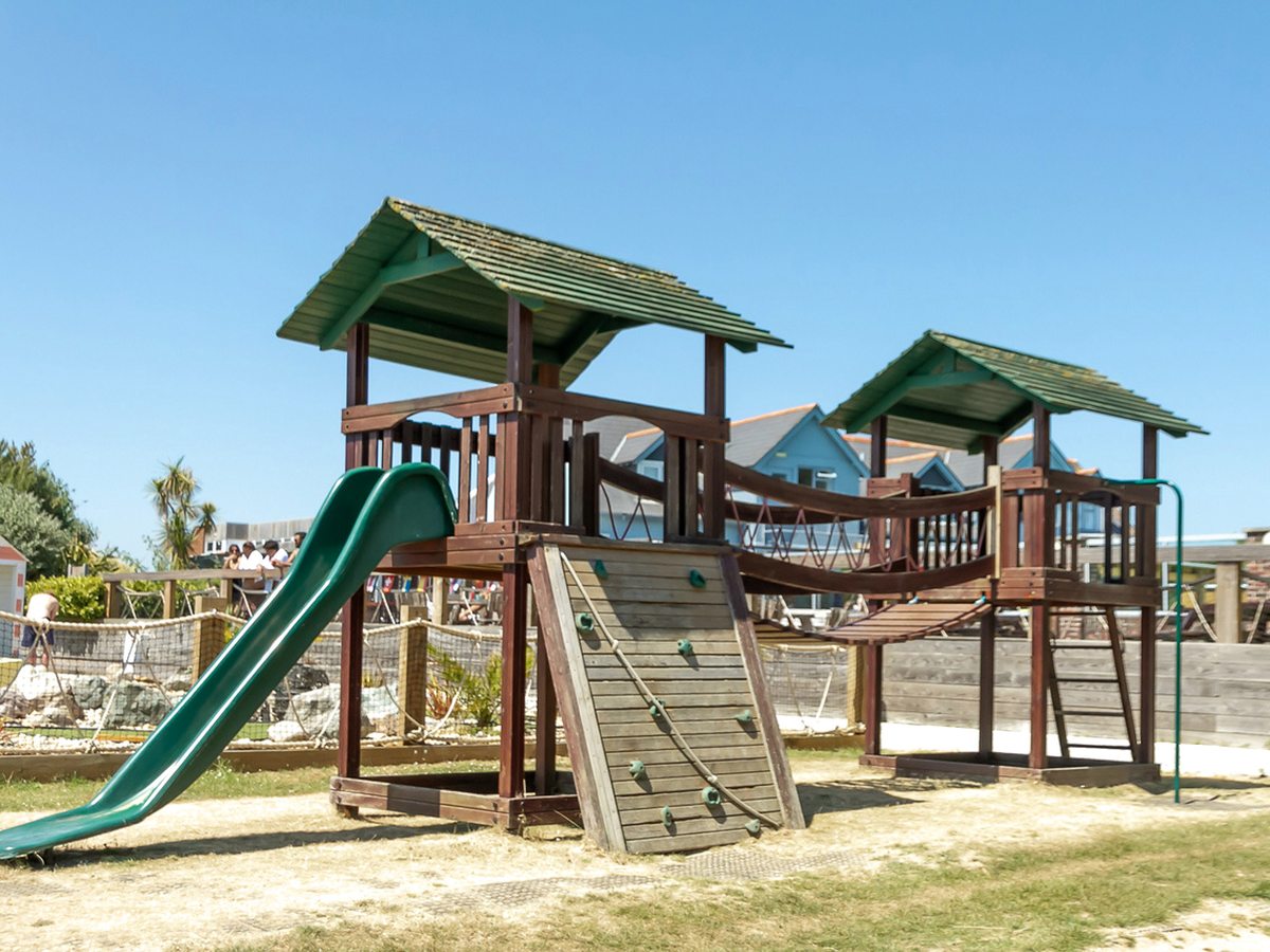 A wooden play frame with two roofed towers, a green slide, a climbing ramp and a rope bridge on dry grass, with blue chalets behind.