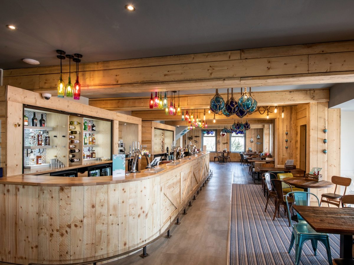 A long curved pine bar with beer taps and bottle shelves, hanging glass-float lights and wooden tables and chairs beyond.
