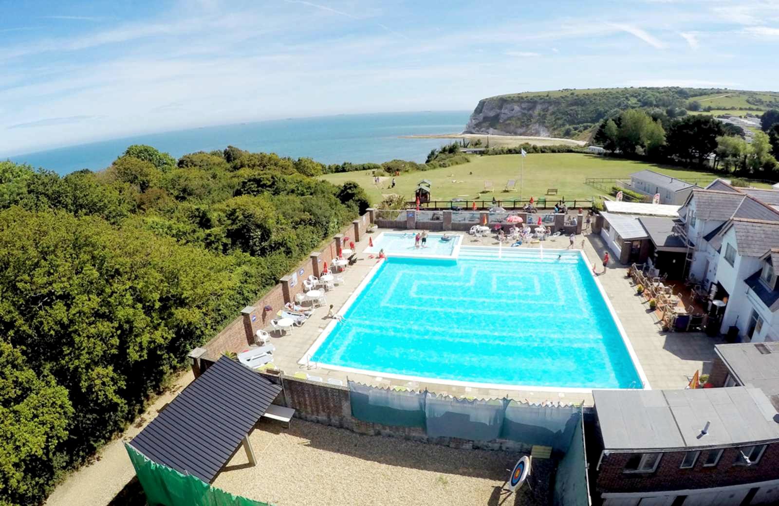 The outdoor pool at Whitecliff Bay Holiday Park on the clifftop, with loungers and tables around it, the bay and a white chalk headland beyond.