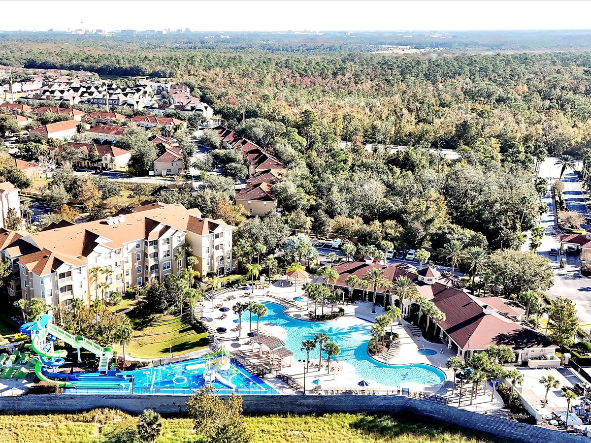 Aerial view of a large resort of low-rise buildings and villas set among trees, with a curved blue swimming pool and colourful water park at its centre.