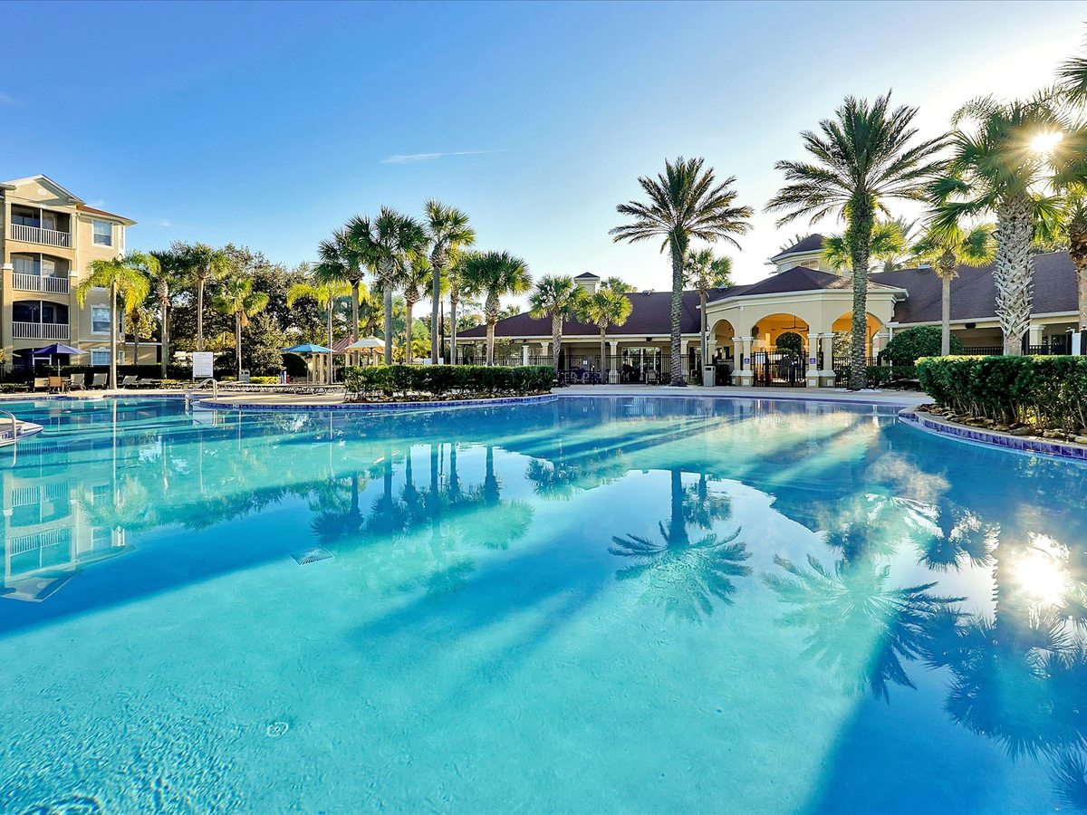 A large turquoise swimming pool with a paved deck, sun loungers, palm trees and an arched clubhouse building behind it.