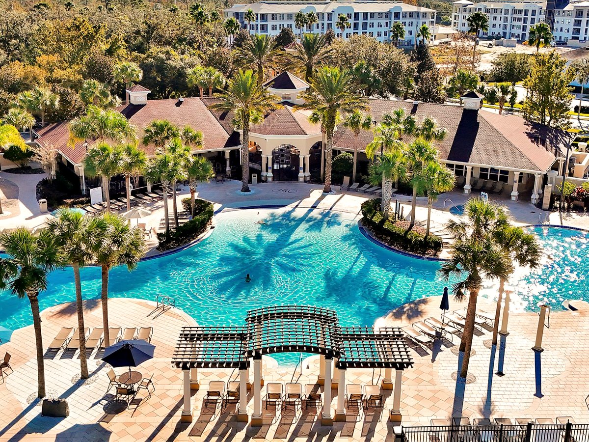 Elevated view of a lagoon pool with a footbridge, poolside tables and chairs, palm trees and the clubhouse behind.