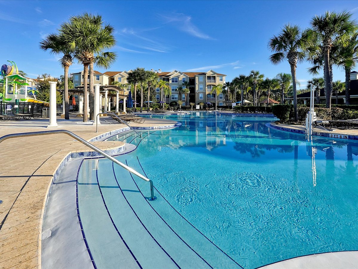 A large blue pool with a broad, gently graduated shallow entry and handrails, edged by palm trees and resort buildings.