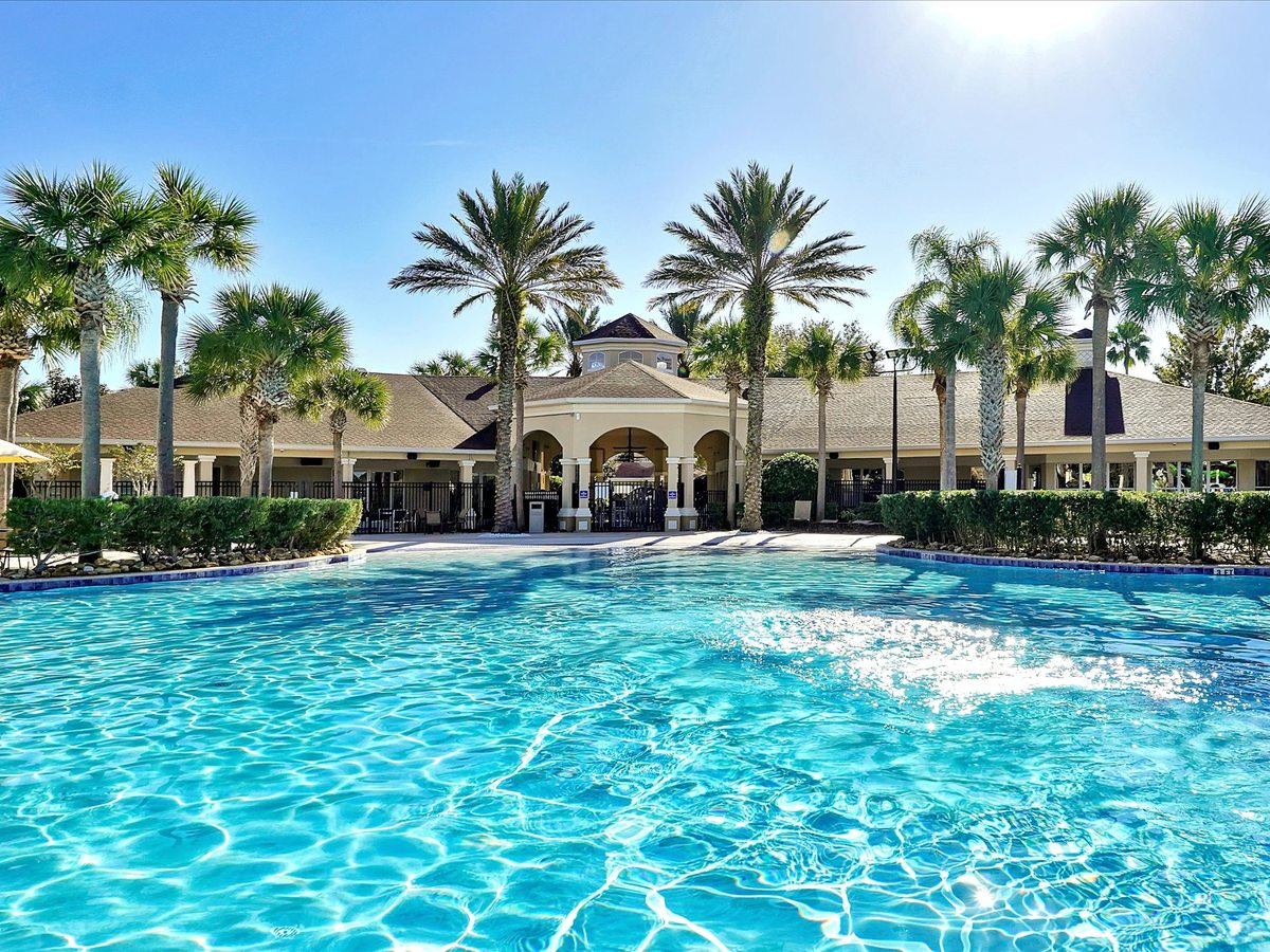 A cream, symmetrical clubhouse with an arched entrance and clipped hedges, seen across a calm blue swimming pool under a clear sky.