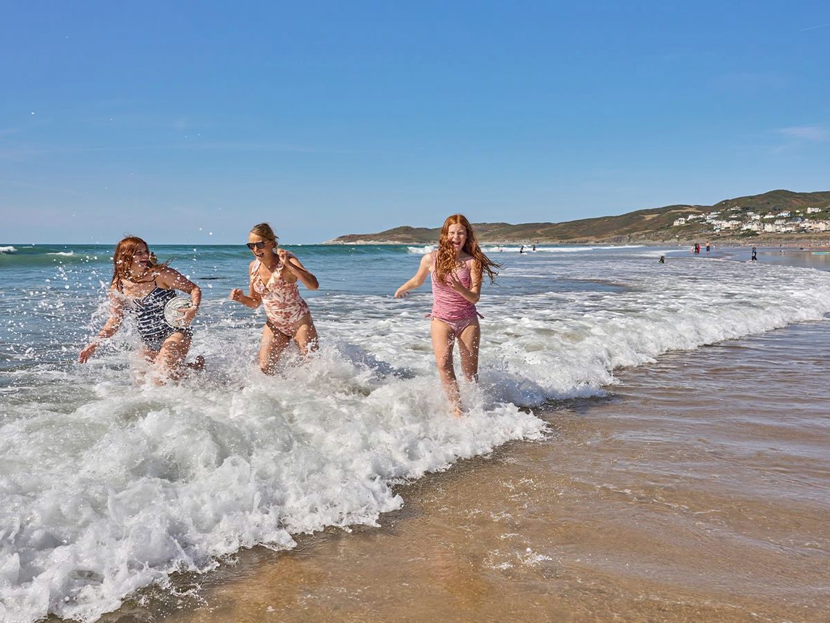 Three girls running through breaking white surf on a wide sandy beach, with a headland and a hillside village in the distance.