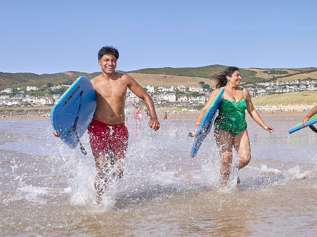 A teenage boy and a woman running out of the shallows carrying bodyboards, with the village of Woolacombe and green headlands behind them.