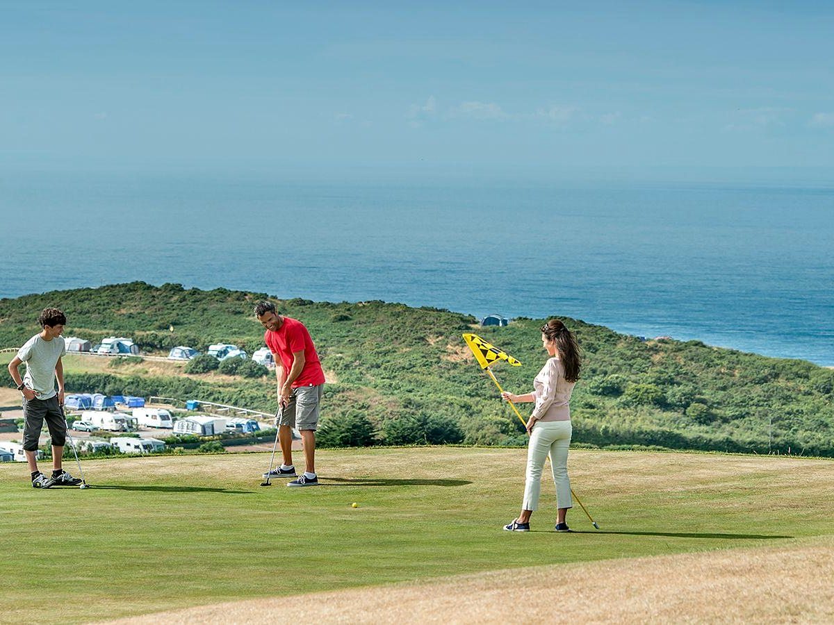Three people putting on a clifftop green, with touring caravans and tents on the slope below and the open sea beyond.
