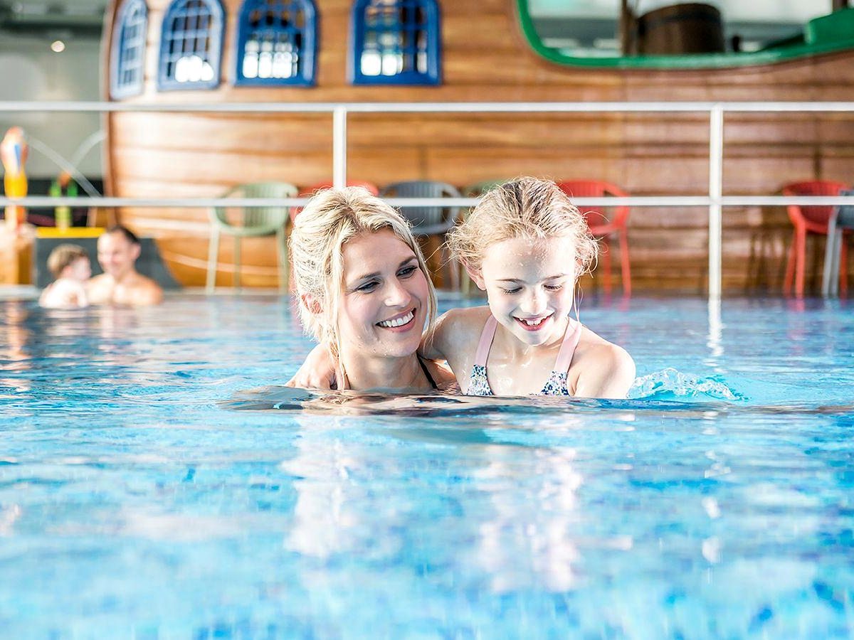 A woman and a girl swimming together in a shallow indoor pool in front of a large wooden pirate-ship structure with painted blue windows.