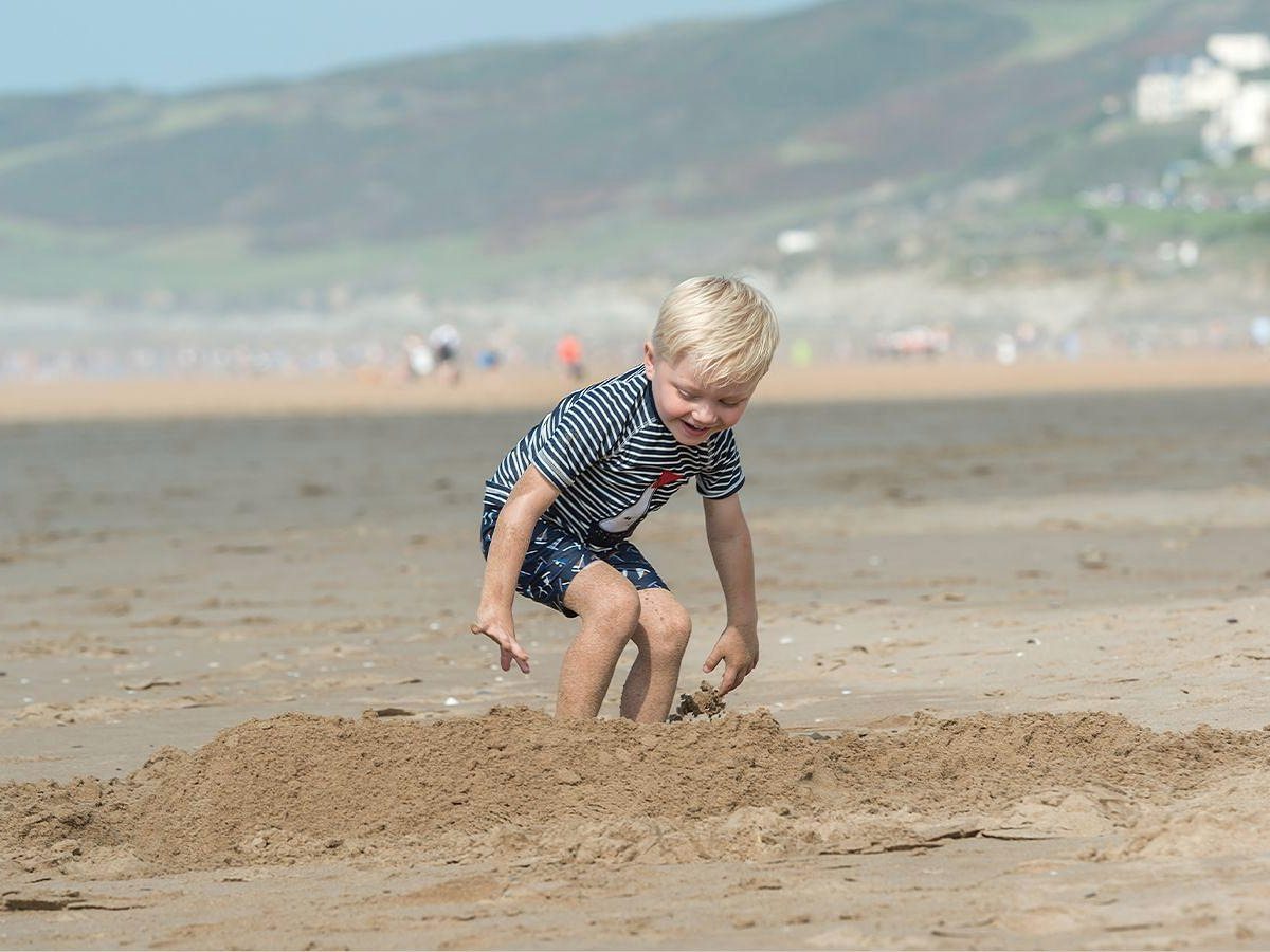 A young boy in a striped rash vest crouching to dig in a mound of wet sand on a wide beach, with dunes and a green hillside blurred behind.