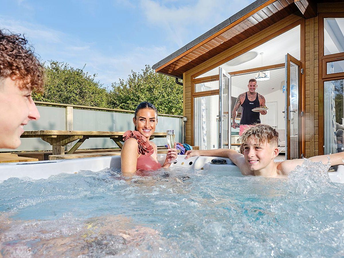 Four people in a bubbling hot tub on a timber deck outside a cedar-clad lodge, with bifold doors folded back and a man carrying a plate of food out from inside.