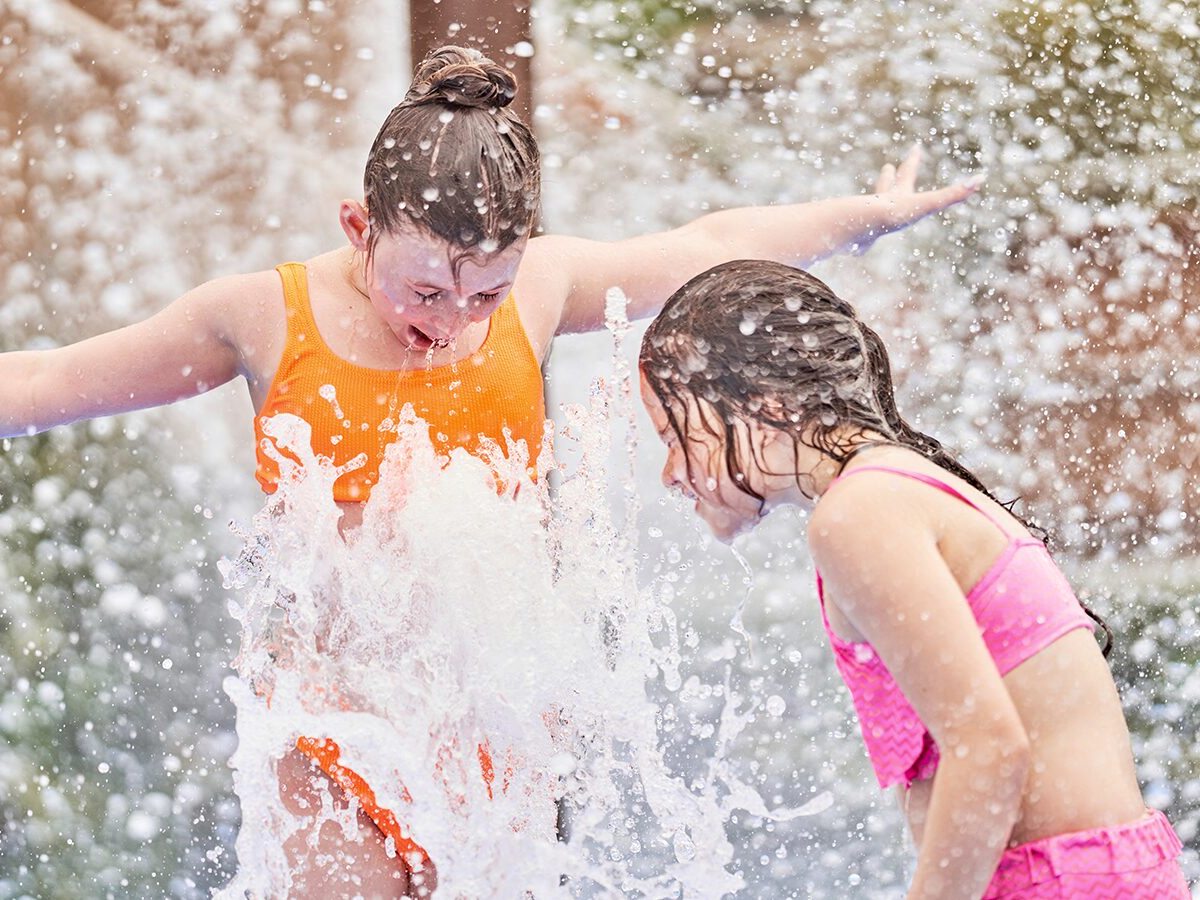 Two girls in swimsuits laughing as water sprays over them outdoors in bright sunshine.