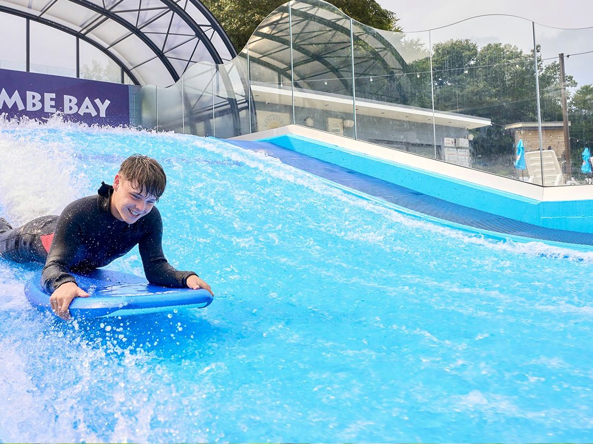 A boy in a wetsuit riding a bodyboard on the fast sheet of blue water of a surf simulator, under a curved glass and steel canopy.