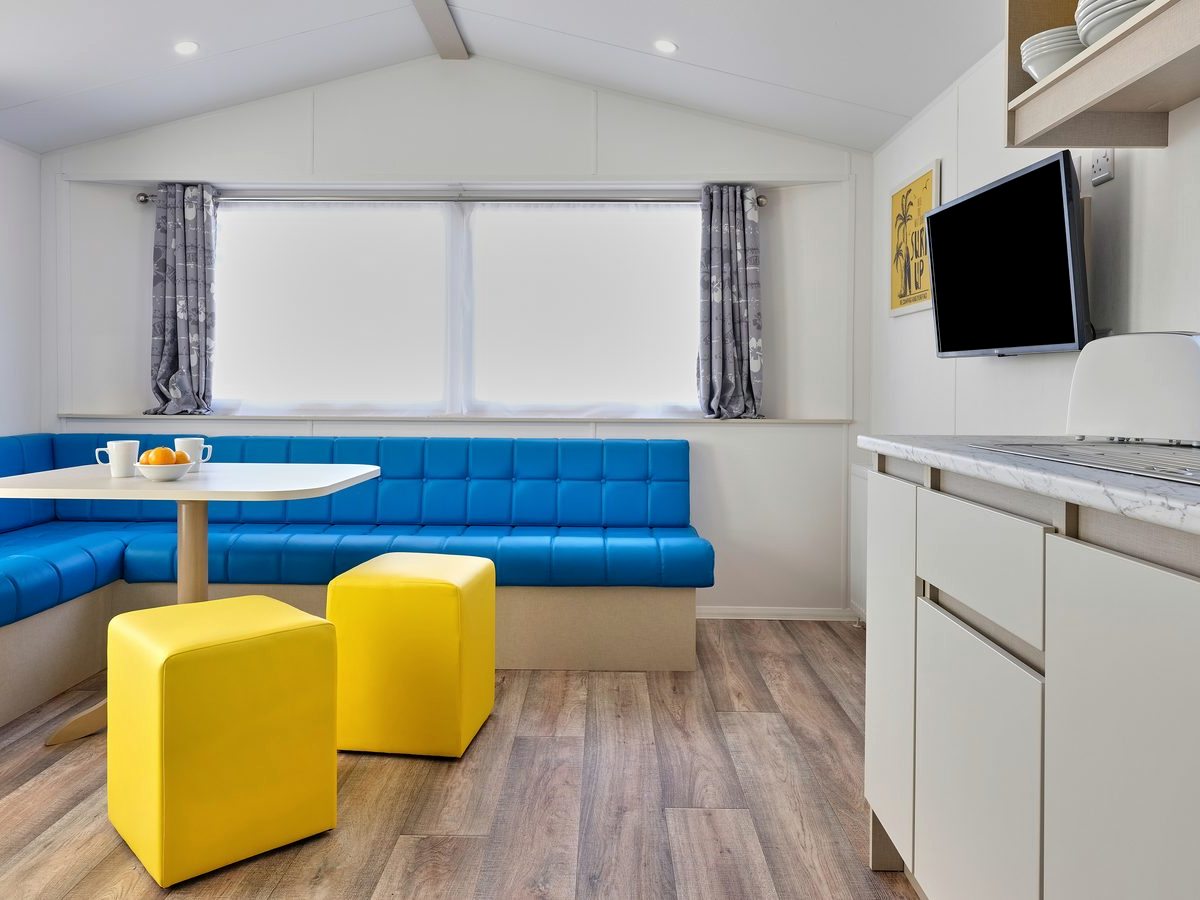 Caravan dining area with a blue wraparound bench seat, a white table, two yellow stools and a wall-mounted television beside white kitchen units.