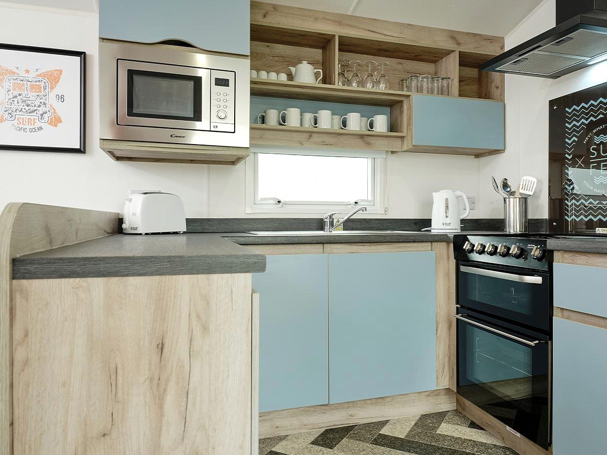 Compact caravan kitchen with pale blue cupboard doors, oak-effect open shelving, a gas cooker and extractor hood, a microwave, a toaster and a kettle.