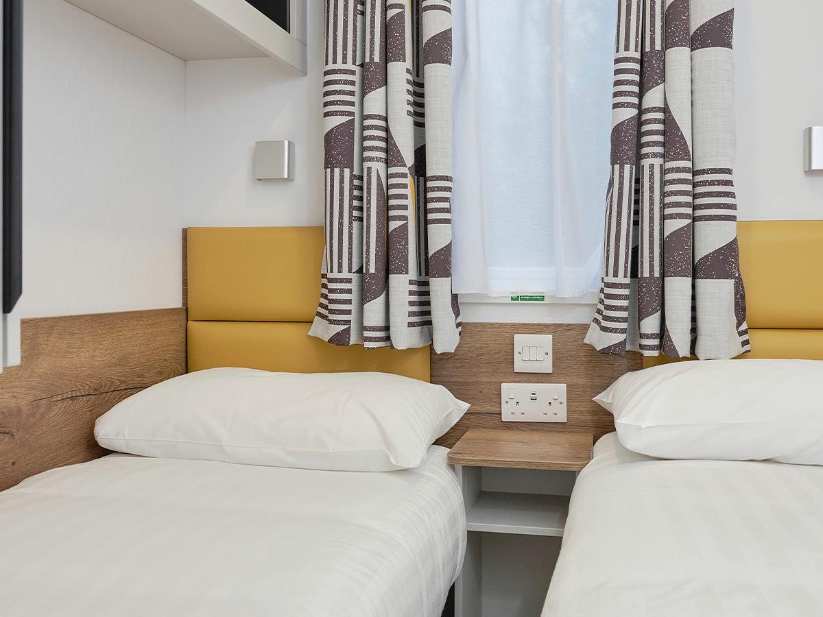 Twin bedroom with two single beds, white bedding, mustard padded headboards, a shared bedside shelf and patterned curtains at the window.