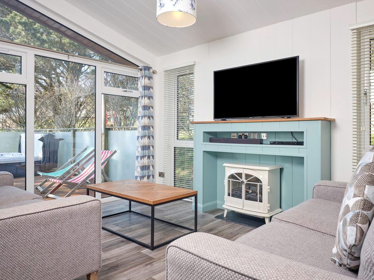 Lodge living room with grey sofas, a wooden coffee table, an electric stove below a wall-mounted television, and glazed doors onto a deck with deckchairs and a hot tub.