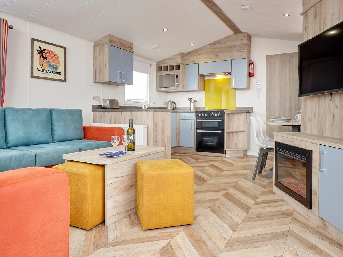 Open-plan caravan living space with a teal sofa, an orange sofa, yellow footstools, an electric fire, a wall-mounted television and a kitchen with a cooker and yellow splashback.