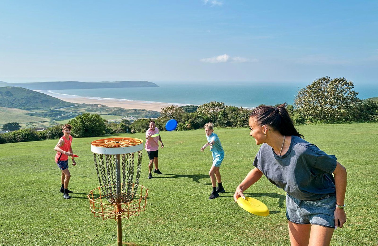 A family throwing discs at an orange disc-golf basket on a clifftop lawn, with the long sweep of Woolacombe Beach and the sea behind them.
