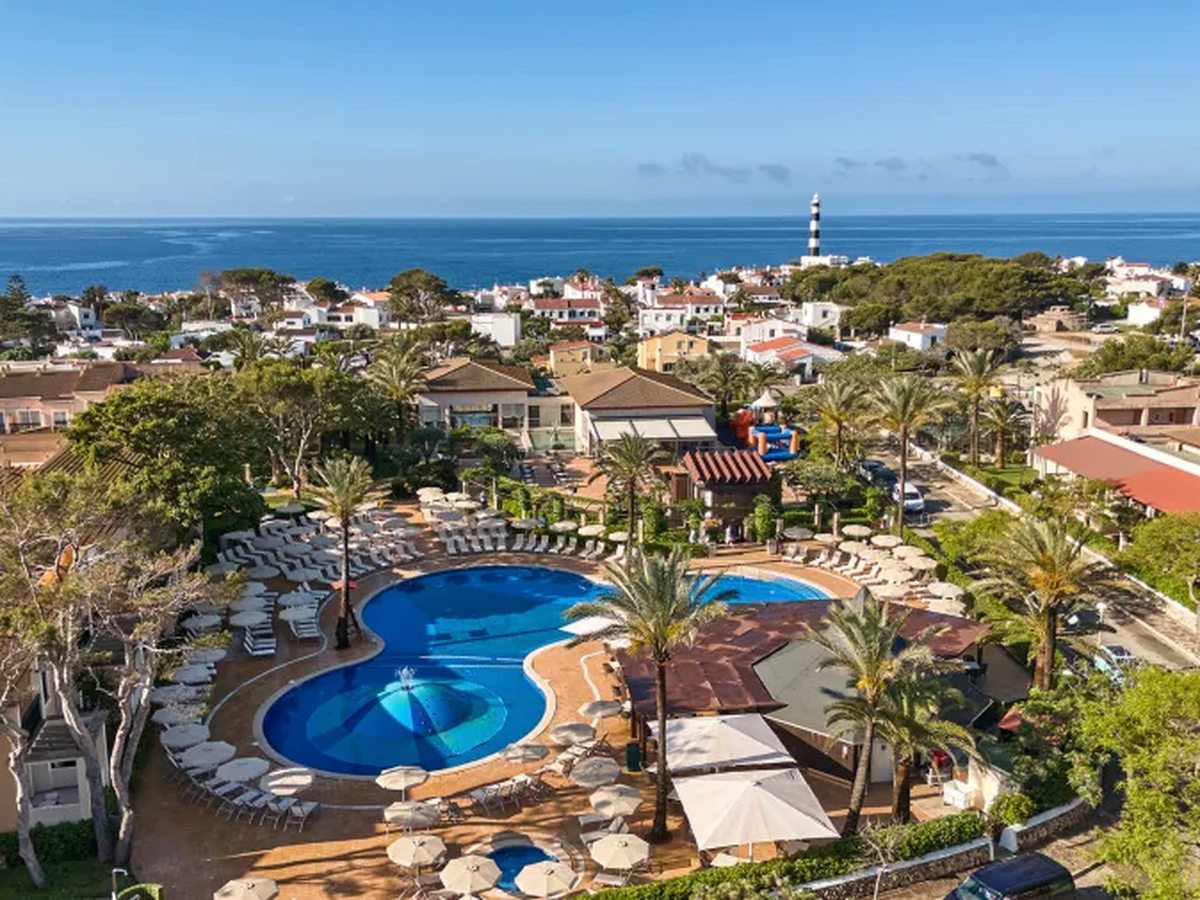 Aerial view of the resort's curved blue pools ringed by sunbeds and palms, with the white houses of Cala'n Bosch, a black and white striped lighthouse and the sea beyond.