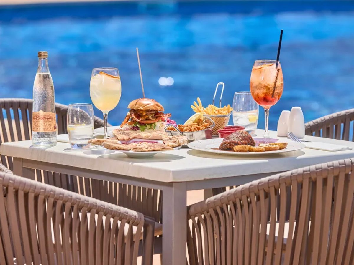 A table beside the pool laid with a burger and chips, flatbreads, a steak sandwich and two cocktails, with woven chairs around it and blue water behind.