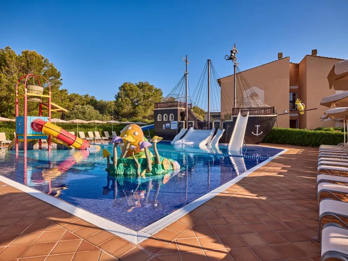 A shallow children's pool with a wooden pirate ship, three white slides, a red tube slide, a tipping bucket and a toadstool play feature.
