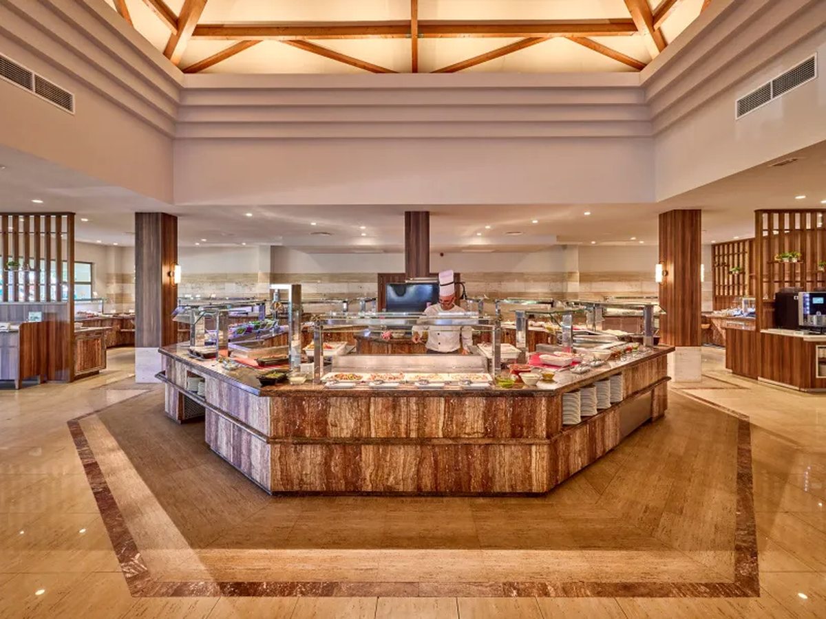 A large buffet island with hot and cold counters under glass screens, stacked plates at one end and a chef working behind it in a high-ceilinged dining hall.