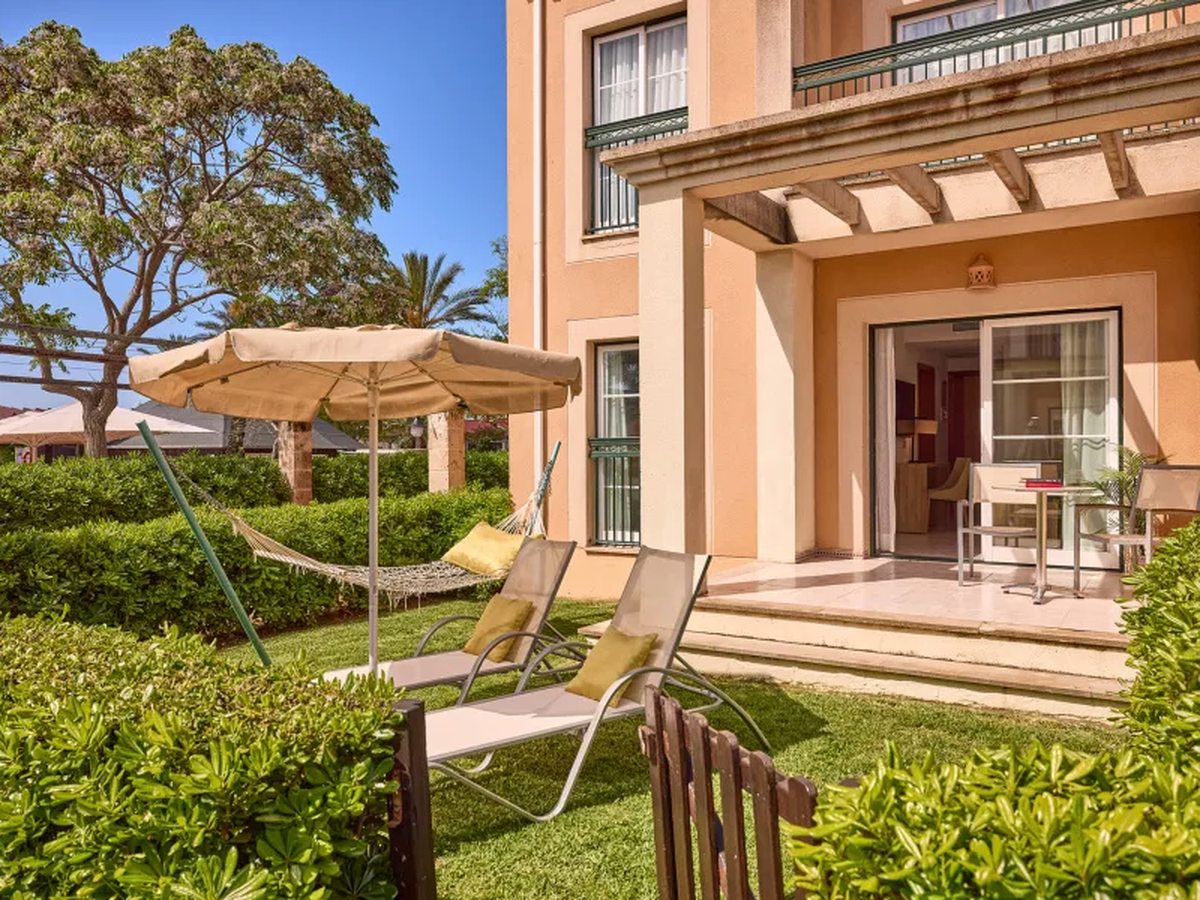 A ground-floor apartment with sliding doors onto a lawned garden with two sun loungers, a parasol and a hammock, enclosed by clipped hedges.