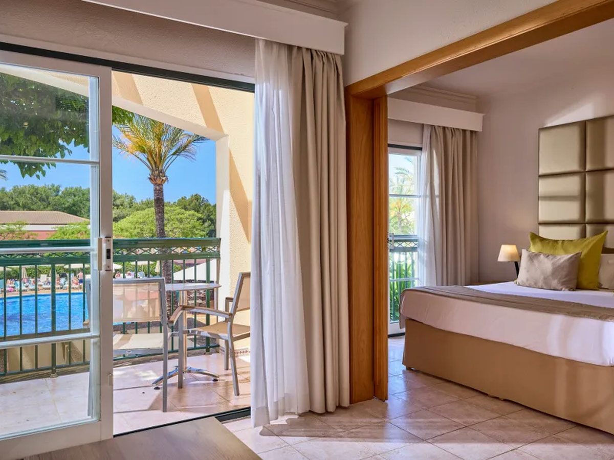 A suite bedroom with a queen bed and padded headboard, alongside a living room with sliding doors onto a balcony with a table and chairs above the pool.